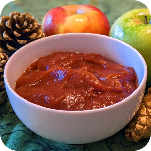 Close-up of homemade apple butter in a bowl with fresh apples and pinecone decorations in the background