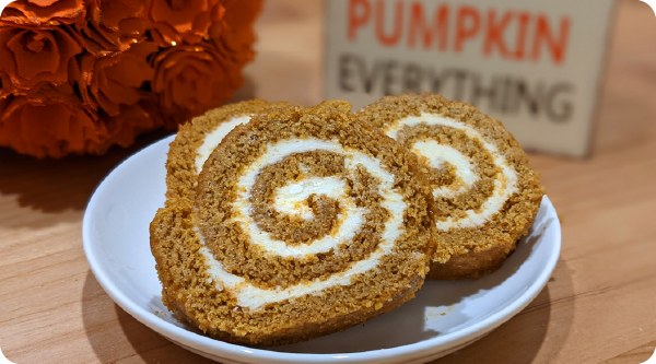 Slices of Pumpkin Cake Roll with cream cheese filling on a plate beside orange flowers and fall decor