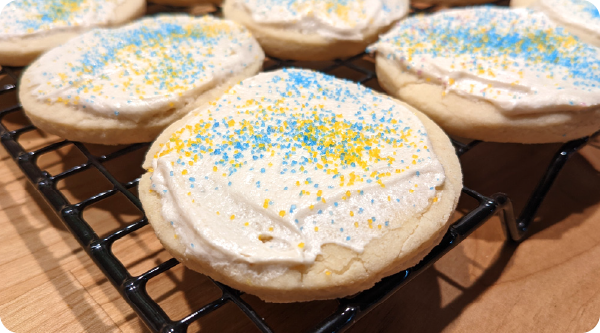 Sour cream cookies topped with white frosting and blue and yellow sprinkles, cooling on a wire rack